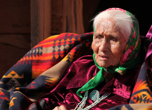 Native American elder woman sitting in a chair with a traditional blanket wrapped around her shoulders