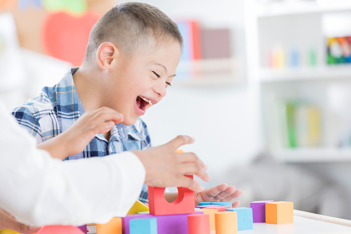 Cheerful preschool age boy enjoys playing with blocks with his teacher. Only the teachers arms are seen in the photo.