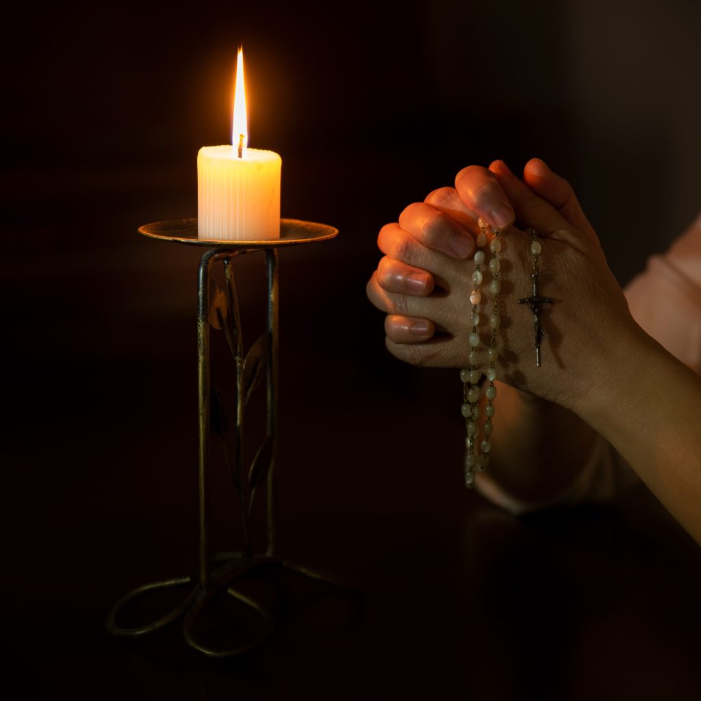 clasped hands holding a hail Mary necklace next to a lit candle